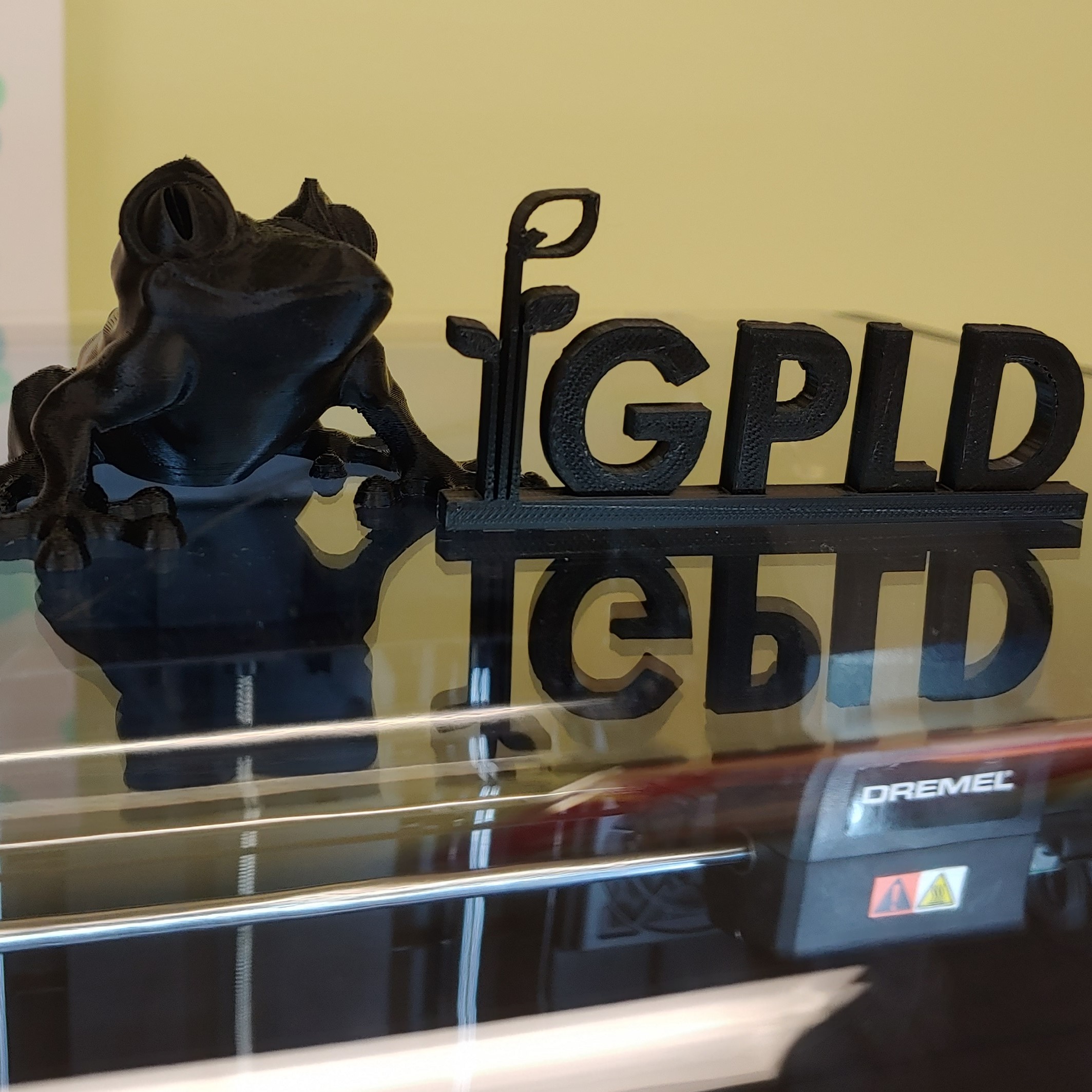 black 3d printed tree frog sitting on top of a dremel 3d printer next to a black 3d printed version of the gpld logo